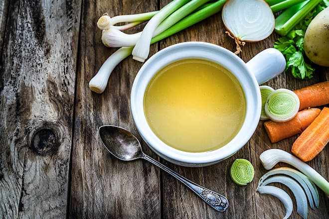 High angle view of a white bowl filled with a healthy vegetables broth shot on rustic wooden table. Fresh multi colored vegetables like carrots, potatoes, celery. parsley and onions are all around the bowl. The composition is at the right of an horizontal frame leaving useful copy space for text and/or logo at the left. High resolution 42Mp studio digital capture taken with SONY A7rII and Zeiss Batis 40mm F2.0 CF lens
