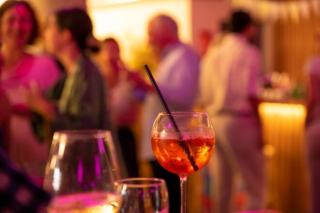 the focus is on a glass with aperol spritz, in the background people standing, out of focus
