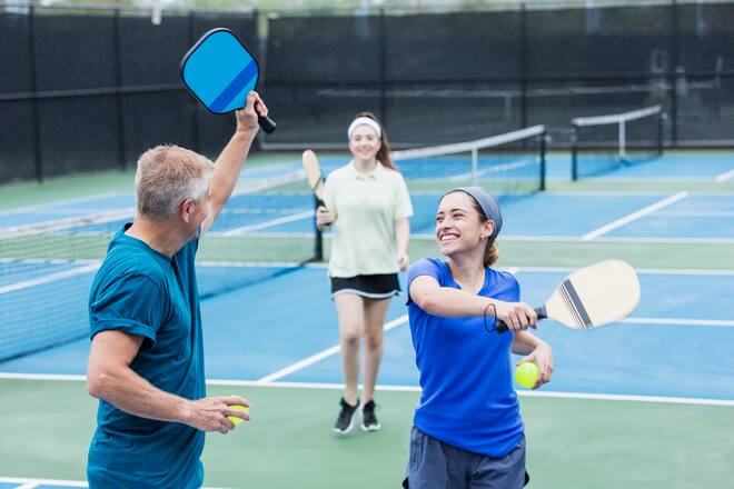 A senior man in his 60s playing pickleball with his daughter, a young mixed race woman in her 20s. She is Hispanic and Caucasian. They are celebrating winning a point, tapping their pickleball paddles together.