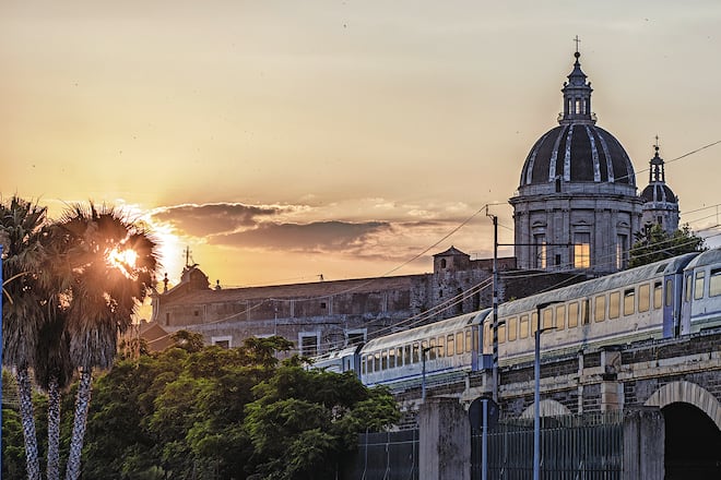Catania, passing the train at sunset over the marina arches. View of the Dome and the city ancient walls