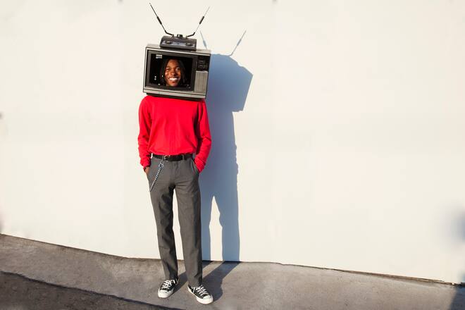 Full length portrait of a smiling young black man with a vintage television set on his head like a helmet looking at camera