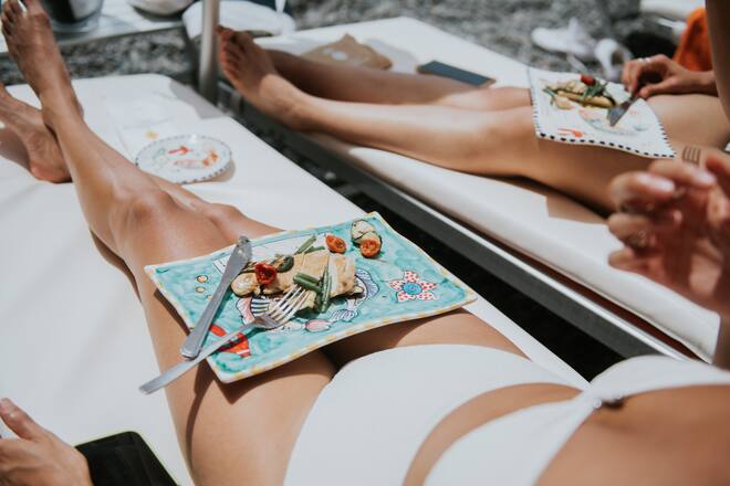 A woman lies on a sun lounger, a plate resting on her thighs. Scene depicts a luxurious holiday dining experience, eating lunch al-fresco, served on a sun lounger. The food looks light, fresh and delicious.