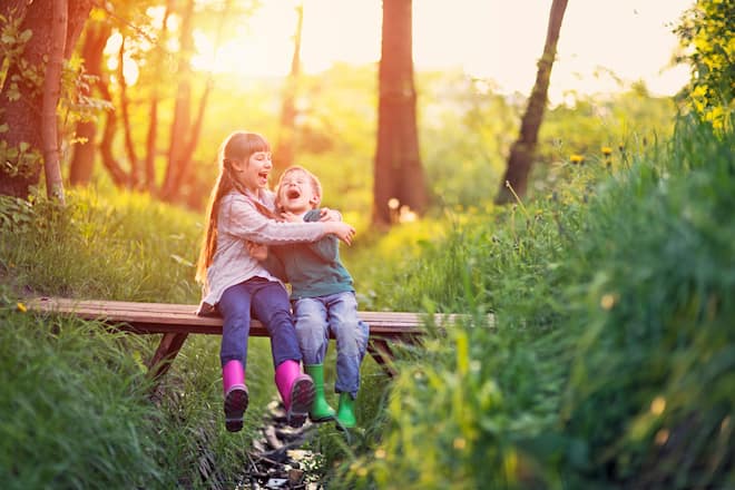 Little boy aged 5 and his elder sister aged 9 are sitting on the small bridge in the forest. They are enjoying beautiful nature and the sunset, laughing happily.