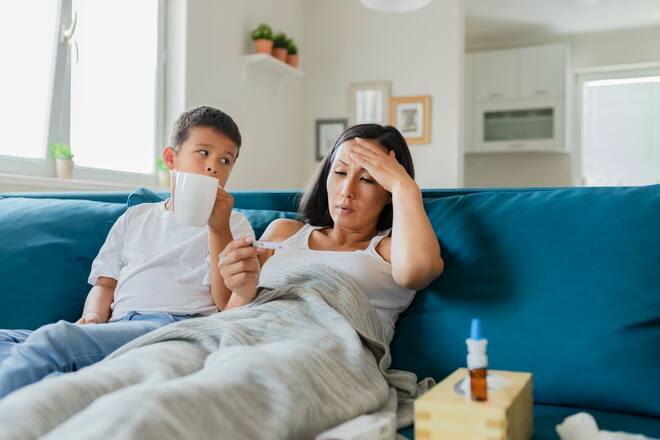 Sick Mother With Thermometer Laying in Bed Covered With a Blanket Checking Temperature While Her Little Son Takes Care of His Mother and Brings Her Tea. Asian Woman Feeling Sick With a Cold and Holding Thermometer With Her Son Who Sits Next to Her and Helping His Mom