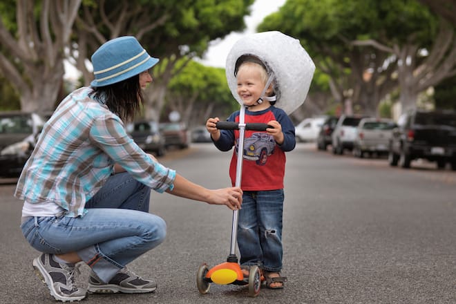 Mother smiling at toddler on scooter wearing helmet wrapped in bubble wrap